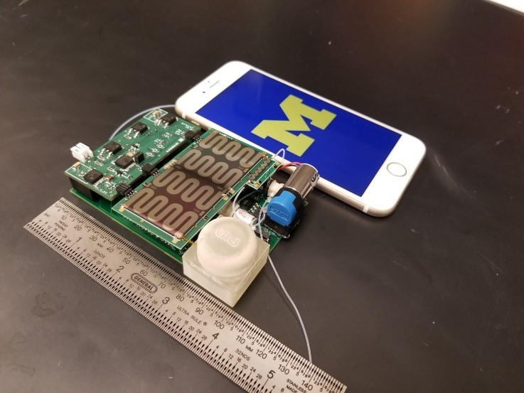 The MAEGLIN sensor, outside its case, with a phone and a ruler for scale. It is about four inches (ten centimeters) square.